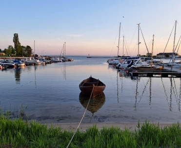 Ł&oacute;dź na pierwszym planie i marina w tle, z odbiciami w wodzie i spokojnym, wieczornym światłem.