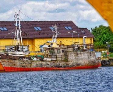 Statek lub kuter przy porcie na tle charakterystycznego budynku &ndash; mocny, kolorystyczny kadr portowy.