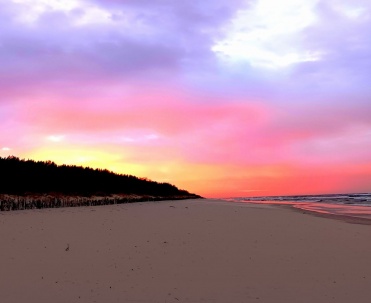 Plaża pod r&oacute;żowo-fioletowym niebem o zmierzchu, z pastelową kolorystyką i miękkim światłem.
