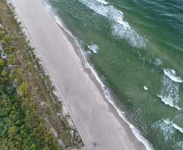 Widok z lotu ptaka na plażę i morze w okolicach Jastarni. Szeroki pas piasku, zieleń i fale Bałtyku pokazują naturalne piękno P&oacute;łwyspu Helskiego oraz wyjątkową przyrodę między Jastarnią a Juratą.